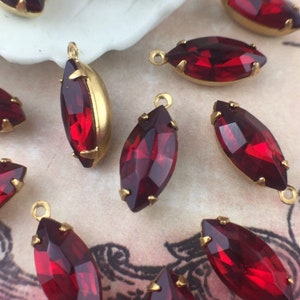 Könnte beinhalten: Marquise-förmige rote Kristallanhänger mit goldfarbenen Metalleinfassungen. Die Kristalle sind facettiert und fangen das Licht ein. Jeder Anhänger hat oben eine kleine Schlaufe zum Befestigen an Schmuck. Der Hintergrund ist eine weiche, neutrale Farbe.