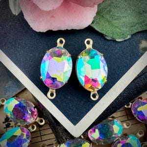 May include: Two oval-shaped iridescent crystal pendants with gold-colored metal settings. The crystals reflect a spectrum of colors, including pink, green, and blue. Additional crystals are scattered around the pendants. The background includes a dark blue surface and floral elements.