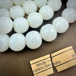 May include: Close-up of a strand of white, marbled beads. The beads are round and have a speckled pattern. A torn tag reads "Made in Western Germany". The beads are on a brown surface.