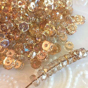 May include: A close-up of a pile of iridescent gold and clear glass beads. The beads are round and have a faceted design. There is a strand of beads in the foreground.