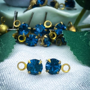 May include: Close-up of several small, round, dark blue crystal stones set in gold-coloured metal. Some have a small loop attached. The crystals are faceted, reflecting light. The background is a soft, blurred mix of green and white.