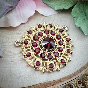 May include: A gold-colored, circular pendant with a central, large, faceted, red gemstone surrounded by smaller red gemstones. The pendant has an ornate, filigree design and two small loops for attachment.