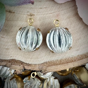 May include: Two clear, round, ribbed glass charms with gold-tone metal findings. The charms have a loop at the top for hanging and are accented with small gold-tone metal prongs. The background is a light brown wooden surface.