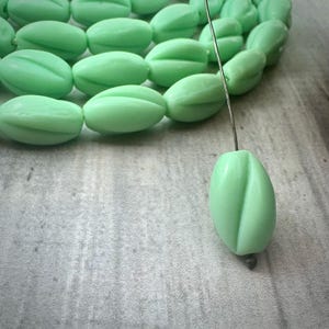 May include: A close-up of light green oval-shaped beads with a groove down the center. The beads are strung on a thin wire, with some beads in focus and others blurred in the background. The beads are on a light gray surface.