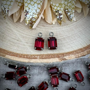 May include: Two red glass emerald-shaped charms with silver settings and loops. The charms are on a wooden surface. There are many more charms in the background.