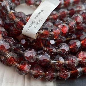 May include: Strand of faceted, dark red beads. The beads are a deep burgundy color with a multifaceted design. A tag reads "Made in Western Germany". The beads are strung together, ready for jewelry making.