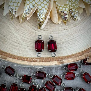 May include: Two red glass rectangular-shaped gems with silver settings and loops on each side. The gems are on a wooden surface. There are many more red gems in the background.
