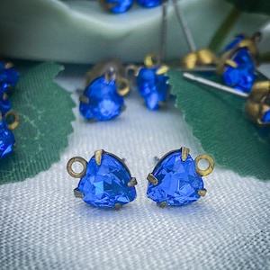 May include: Two blue triangle-shaped glass rhinestones with gold metal settings and loops. The rhinestones are on a white fabric background.