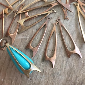 May include: An assortment of vintage-style jewelry components. The image features several copper-toned metal teardrop-shaped pendants and a turquoise and gold-toned pendant. The components are scattered on a textured, neutral-toned surface.