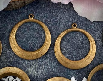 May include: Close-up of several antique brass-colored metal pendant hoops. Each hoop has a circular design with a flat bottom and a small loop at the top for attaching to a chain or other jewelry component. The hoops are arranged on a dark blue textured surface.