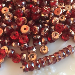 May include: A pile of red faceted glass beads with a gold metallic finish. The beads are arranged in a loose pile, with some beads spilling out of the pile. The beads are all the same size and shape, and they have a shiny, reflective surface.