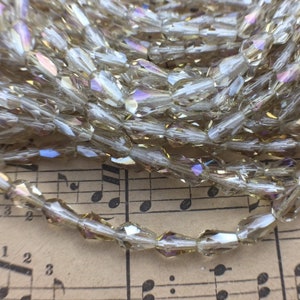 May include: A strand of iridescent glass beads in a light gold color. The beads are faceted and have a teardrop shape. The beads are arranged in a pile on a sheet of music.