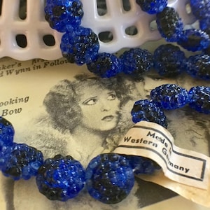 以下が含まれることがあります： ファセット加工された青と黒のビーズが付いたヴィンテージネックレスのクローズアップ。ビーズはつながっており、「Made in Western Germany」と書かれたタグが付いています。ネックレスはヴィンテージ写真に表示されています。