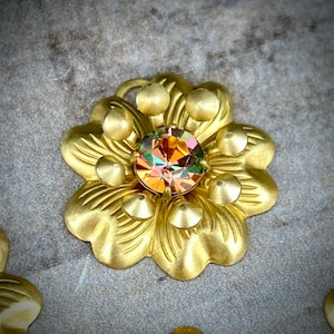 May include: A gold-toned metal flower charm with a faceted iridescent gemstone in the center. The flower has seven petals and six round accents around the gemstone.
