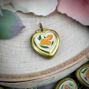 Può includere: Un medaglione a forma di cuore, color oro, con un motivo floreale arancione su sfondo bianco. Il fiore ha foglie verdi. Il medaglione è aperto. È posto su una superficie di legno.