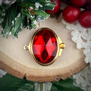 Pode incluir: Uma gema vermelha ovalada e facetada, emoldurada por uma estrutura de metal dourado. A gema tem um corte brilhante, refletindo a luz. A moldura inclui uma pequena argola e um fecho. A peça é exibida sobre uma superfície de madeira.