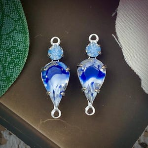 May include: Two teardrop-shaped, blue and white glass pendants with silver-colored metal settings. Each pendant has a small, round, light blue stone at the top and a loop for hanging. The blue glass has a swirled pattern.