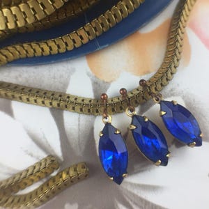 May include: Three blue teardrop-shaped crystal earrings dangle from gold chain links. The earrings are on a white background with a gold chain in the background.