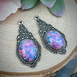 May include: Two silver-toned teardrop-shaped pendants with iridescent, oval-shaped centers. The centers display a mix of pink, purple, and blue hues. The pendants have a decorative border and a small loop at the top for hanging. The background is a wooden surface.