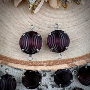 Può includere: Due cabochon in vetro viola scuro con montature e anelli in metallo argentato. I cabochon hanno una texture a coste.
