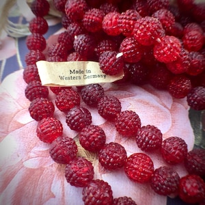 Può includere: Primo piano di una collana di perline di vetro rosso vintage. Le perline sono di un rosso intenso e traslucido con una superficie strutturata, simile a una lampone. Un'etichetta recita "Made in Western Germany". Le perline sono disposte su un tessuto a motivi floreali.