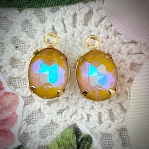 May include: A pair of oval iridescent crystal pendants with gold-coloured metal settings. The crystals have a yellow-orange hue with flashes of blue and purple. Each pendant has a small loop at the top for attaching to a chain or other jewellery component.