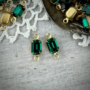 May include: Two emerald green rectangular glass stones with gold metal connector loops. The stones are set in a gold metal frame.