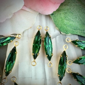 May include: Close-up of several green marquise-shaped crystal pendants with gold-colored metal settings and loops. The crystals have a faceted design, reflecting light. The pendants are arranged on a white surface, with a blurred floral background.