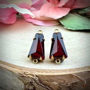 May include: A pair of faceted, dark red gemstone earrings with gold-colored metal accents. The earrings have a geometric, triangular shape with a small loop at the top for hanging. The gemstones are set in a gold-colored metal base.