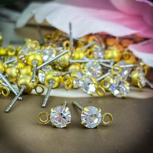 May include: A close-up of a pile of gold-toned earring findings with clear crystal rhinestones. The findings have a loop at the top and a post with a small ball at the end. The rhinestones are round and have a faceted surface.