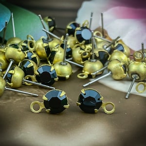 Könnte beinhalten: Nahaufnahme von schwarzen Strass-Ohrring-Findings mit goldfarbenen Fassungen. Die Findings sind auf einer braunen Oberfläche verstreut.