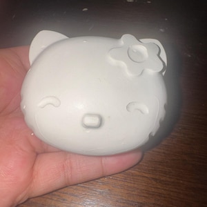 May include: A white, round, Hello Kitty-shaped object with a flower on its head. The object has small, curved lines for eyes and a rectangular nose. The object is held in a hand.