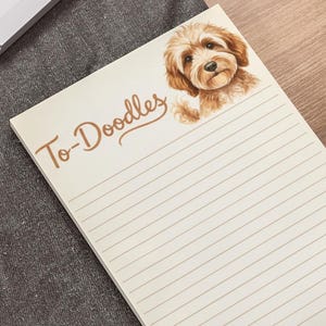 May include: Cream-colored notepad with horizontal lines and the words "To-Doodles" in brown script. A watercolor illustration of a brown and tan Doodle dog is on the top right. Paw prints are in the bottom right corner. The notepad is on a desk.