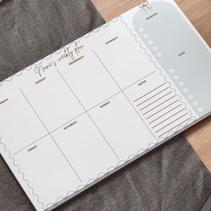 May include: A white weekly planner with a light blue border and grid lines. The planner includes sections for each day of the week, priorities, to-do list, and notes. The text "Name's weekly plan" is written in cursive.
