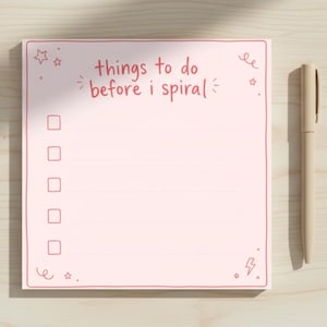Może przedstawiać: Różowy notatnik z napisem "things to do before I spiral" w stylu odręcznym. Notatnik ma jasnoróżową obwódkę i pola wyboru. Beżowy długopis spoczywa obok notatnika na jasnej drewnianej powierzchni.