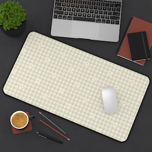 May include: A rectangular mouse pad with a black border features a light green and white gingham pattern with small white daisies. A white computer mouse sits on the pad, with a laptop, notebooks, and a cup of coffee nearby.