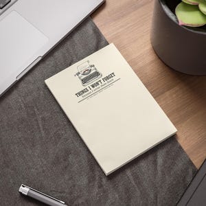Può includere: Un blocco note color crema con un'illustrazione in bianco e nero di una macchina da scrivere vintage nella parte superiore. Sotto la macchina da scrivere, il testo recita: THINGS I WON'T FORGET because I wrote them down. Design rettangolare.