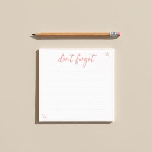 Op de afbeelding: Een witte notitieblok met de roze tekst "don't forget", vergezeld van een houten potlood met een roze gum. Het notitieblok heeft gelinieerde pagina's en sterillustraties.