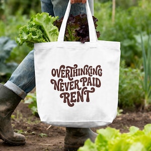 May include: A white canvas tote bag featuring the dark brown text "OVERTHINKING NEVER PAID RENT" in a bold, wavy, retro-inspired font. The bag has two sturdy white shoulder straps and is filled with fresh green and purple leafy vegetables.