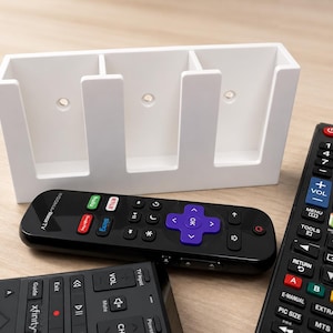 May include: A white plastic remote control holder with three vertical slots and pre-drilled mounting holes, positioned behind three black television remote controls with various buttons, including a purple directional pad and coloured function keys.