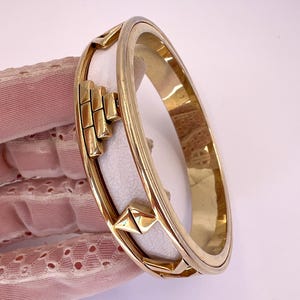 Puede incluir: Un brazalete dorado con una inserción de cuero blanco. El brazalete presenta diseños geométricos y una forma rectangular. El brazalete tiene aproximadamente 6,35 cm de diámetro.
