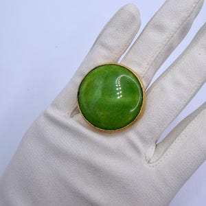 Könnte beinhalten: Ein großer, runder Ring mit einem glatten, leuchtend grünen Stein, der in eine polierte, goldfarbene Metallfassung eingefasst ist. Der Stein hat eine glänzende, reflektierende Oberfläche mit subtilen Variationen im Grünton für ein einheitliches Aussehen.