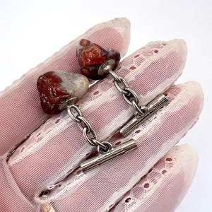 Puede incluir: Gemelos con cabujones de piedra natural en tonos rojos, marrones y blancos. Las piedras están conectadas por una cadena de plata y un cierre de barra. Los gemelos se muestran sobre un fondo blanco.
