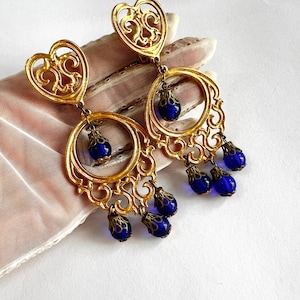 May include: Ornate gold-coloured earrings with a heart-shaped top and a circular design. Each earring has dangling, teardrop-shaped, deep blue beads. The earrings feature an intricate, filigree-style design, adding a vintage aesthetic.