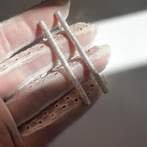 Può includere: Un paio di orecchini a barra d'argento con superficie strutturata. Gli orecchini sono dritti e sottili, riflettendo la luce. I gioielli sono esposti su un tessuto rosa e di pizzo.