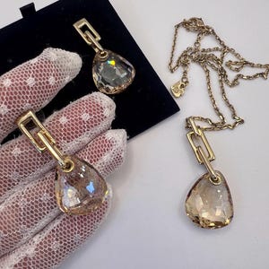 Pode incluir: Conjunto de joias em tom dourado com um colar com pingente e brincos a condizer. Os pingentes e brincos têm um cristal facetado em forma de gota com um elo retangular dourado. Os cristais têm um tom castanho claro.