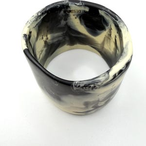 Könnte beinhalten: Ein breites, zylindrisches Armband mit Marmormuster. Das Armband hat eine Mischung aus schwarzen, cremefarbenen und weißen Farben, die ein wirbelndes Muster erzeugen. Das Wort "DESIGN" ist auf der Oberkante sichtbar. Dieses Accessoire ist für den Alltag geeignet.
