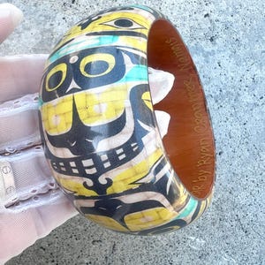 May include: A wooden bangle bracelet with a Native American design. The bracelet features yellow, black, and turquoise colours. The interior is a warm brown. The text "by Ryan Cranmer Namgis" is visible on the inside of the bracelet.