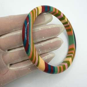 May include: A circular bangle bracelet featuring a repeating pattern of vertical stripes in shades of peach, sage green, dark red, and teal. The surface of the bangle is smooth and glossy, with a consistent thickness throughout the entire round shape.