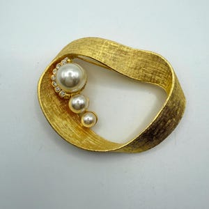 May include: A gold-toned brooch featuring a textured, ribbon-like oval frame. The design includes three graduated white faux pearls arranged in a vertical line, with the largest pearl accented by a semi-circle of small, clear, sparkling rhinestones.